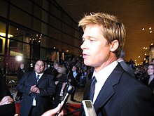 A male with dyed blonde hair is being interviewed. He is wearing a black suit and tie, with a white shirt, and is standing on a red carpet. People standing behind barricades are visible in the background, while microphones are visible in the foreground.