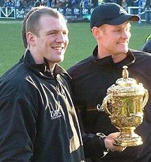 2 hommes, vêtus en noir, l'un portant une casquette. Ils portent une coupe.