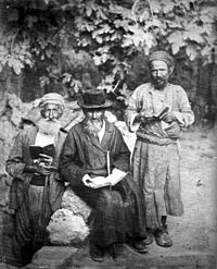 A sepia photograph shows three elderly Jewish men sporting beards and holding open books, posing for the camera. Against a backdrop of leafy vegetation, the man in the centre sits, wearing a black hat and caftan, while the two others stand, wearing lighter clothes and turbans.