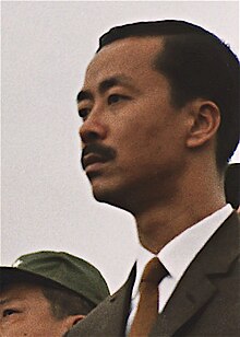 A middle-aged man with side-parted black hair and a moustache, in a black suit, white shirt and brown tie. To the left is a clean-shaven Asian man with black hair and a green military cap.