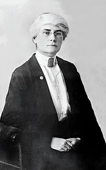 A white woman with white hair and dark eyebrows, wearing eyeglasses, a high-collared white blouse with many fabric-covered buttons down the front, and a dark jacket