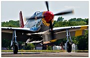 A single nosed-propeller plane is on the ground on its wheels with the propeller in motion. The plane is viewed from the front, but the red nose faces slightly to the right. The plane has black propellers. Parts of the wings and propellers are crpped from view.