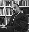 Ralph Ellison is pictured sitting in a chair before a bookcase. He is wearing a suit and has a mustache and receding hair-line.