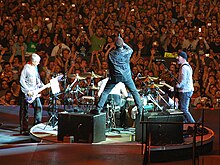 U2 performs on a stage. Bono stands in the center facing away towards a drum kit, with each leg on an amplifier. The Edge, playing guitar to his right, faces him. Adam Clayton, playing bass to his Bono's left, faces him. Fans are visible in the background