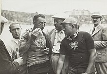 Photographie d'un cycliste s'essuyant le front, entouré de plusieurs hommes.