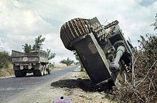 Photographie en couleur montrant un char renversé sur le flanc sur le bord d’une route sur laquelle circule un camion.