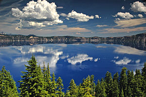 Photograph of Crater Lake depicting clear blue waters reflecting fluffy clouds with forestry peeking out in the forefront