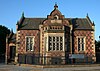 Photograph of a red-brick building with blue-brick decoration, with a central shaped gable including a bay window with a stone balustrade above