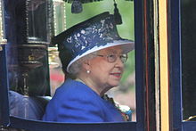 Elizabeth, in formal dress, holds a pair of spectacles to her mouth in a thoughtful pose