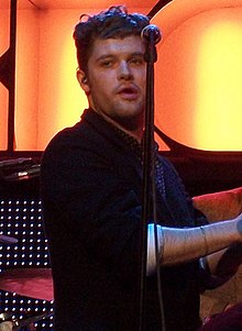 A thirty-six year-old man is shown in three-quarter view. He is standing centre-stage, half-turned with hands clapped together. He is partly obscured by a microphone stand in front. Behind him is a drummer at his kit. Beyond that is the lighting, which includes the partly spelled letters R-O-C-K-C-O-R in large orange-yellow.