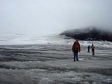 Three climbers standing on a dark glacier with a black mountain in the right background.