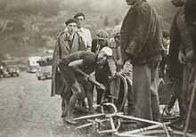 Photographie en noir et blanc montrant un cycliste regonflant son pneu, entouré par quelques hommes.