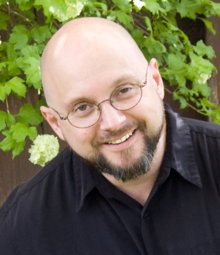 Picture of Howard Tayler wearing a medium blue denim button-up collared shirt. He is looking over the top of his glasses while grinning at the camera.