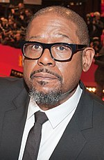 Portrait of an African-American male facing toward his right shoulder. He is wearing a black suit and an unbuttoned white collar shirt.