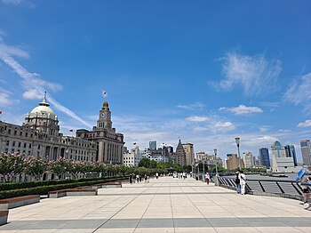 A wide curving walkway along a body of water to the right, crowded with many people and some large orange umbrellas underneath a sky colored by sunset. To the left of the walkway are light brownish-grey stone buildings in various late-19th and early-20th-century architectural styles, with taller buildings in the distance.