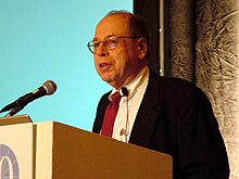 Chest high portrait of man in his sixties in a suit and tie speaking at a microphone in front of a blank blue slide projection