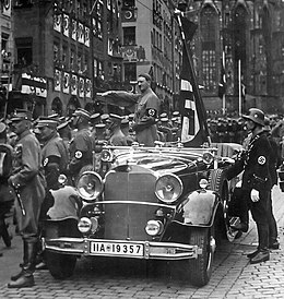 Photographie en noir et blanc de Hitler lors d'un congrès du parti nazi à Nuremberg en 1935. Debout dans une voiture décapotable, il fait le salut nazi à ses partisans qui défilent