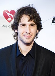 A young man with medium-length wavy hair, smiling slightly and dressed in black business attired.