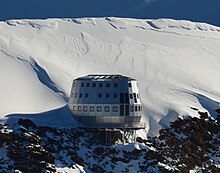 Refuge ovoïde et métallique à la base d'une arête neigeuse.