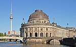 An ornate grey stone building on the point of an urbanized island. The building is connected by two bridges to the neighboring banks