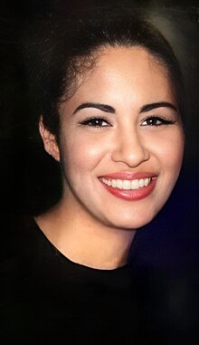 Selena é uma mulher jovem, com cabelos cacheados volumosos. Ela está com roupas brancas e exibe um sorriso para a foto.