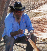 Aboriginal man sitting with black brimmed hat, holding a traditional tool