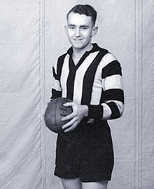 A dark-haired footballer in a long-sleeve black-and-white vertically striped guernsey and black shorts, holding a football