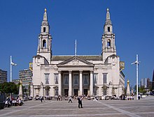 A pedestrian square in sunny weather. At the rear is a large neo-Georgian building of Portland stone. It has a giant portico with four Corinthian columns and a pediment. On the ground floor, there are three entrance doors under the portico and two windows on either side of it. All the doors and windows have Gibbs surrounds and pediments. The window pediments are normal, while those over the doors are segmental. The first floor has seven plain windows, three of them behind the portico, and all of them larger than those on the ground floor. On either side of the building are two tall towers in Wren style with elaborate columned pavilions surmounted by smaller pedimented ones, and on top of each is an obelisk surmounted by a golden owl. In front of the building on each side there are similar obelisks and owls, and prominent gold clocks are attached to the sides of the building at second-floor level. In the background on both sides are tall modern buildings.