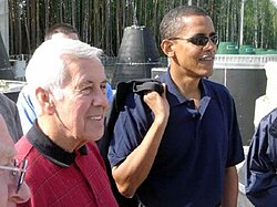 Gray-haired man and Obama stand, wearing casual polo shirts. Obama wears sunglasses and holds something slung over his right shoulder.