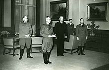 Photographie d'un bureau relativement dépouillé. Quisling, debout et en costume, se tient aux côtés de plusieurs hommes en uniforme dont un a des feuilles dans les mains