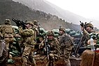 Seven soldiers in beige tactical gear huddle behind a row of green sandbags on a mountainside, pointing rifles in various directions