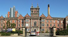 A long mid-Victorian Jacobethan-style building of red brick can be seen beyond a wide entrance with open metal gates and adjacent railings. A car is about to go through the entry barrier. The building has three storeys with, in the centre, a large round-headed window occupying the two storeys over the entrance and a tower above with turrets at the corners. On either side of the tower, bay windows project forward on all three floors, and parapets and six shaped gables can be seen along the roof-line. Behind and to the left can be seen a tall modern building, while behind and to the right there is a tall chimney.