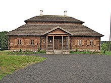 Photographie d'une maison en bois avec un toit en chaume. Un porche et quatre fenêtres se trouvent sur la façade avant