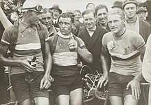 Photographie en noir et blanc montrant trois cyclistes se tenant debout à côté de leur machine, après l'arrivée, deux d'entre eux ayant une bouteille à la main.