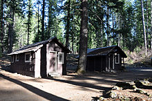 Two small cabins in the woods face a dirt road.