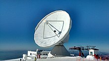 Photo d'une grande antenne au sommet d'une montagne enneigée, à côté de remontées mécaniques.