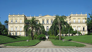 Photograph of an immense off-white building behind two rows of palm trees leading to the entrance, a clear blue sky above