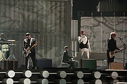 Several men in suits performing on a stage in front of a grey backdrop.