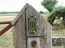 A close-up view of a large wooden post with a pointed top. Affixed to this is a panel containing a yellow outline of an owl and the letters "LCW". Attached to the post are two rusty metal rails to the left and part of a wooden fence with barbed-wire to the right. In the background are several fields, with a low hill far left. To the right is a nearby tree and there is a larger one some distance behind it.