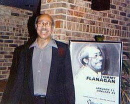 un homme Noir, cheveux gris et lunettes qui pose en souriant devant une affiche