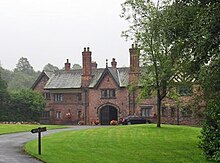 A two-storey house including an archway under a gable and another gable with half-timbering. In front of it is an area of grass and trees.