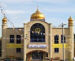 A large symmetrical two-storey building of yellow brick. The centre bay, incorporating the entrance, juts out. It has a large window with a semicircular top on the first floor and above is a golden onion dome on a blue base. At the ends of the frontage are hexagonal pilasters with small octagonal windowed pavilions and onion domes on top. Above the entrance is a white sign saying "The Sikh Temple" in blue. Each side has one two-storey and one one-storey window, and also yellow banners alongside the Temple's name. On each side of the entrance is a wooden seat, and strings of bunting are stretched across the scene.