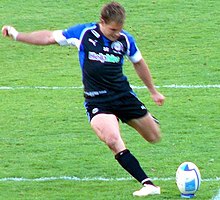 Joueur de rugby, de face, en train de frapper un ballon de son pied gauche.