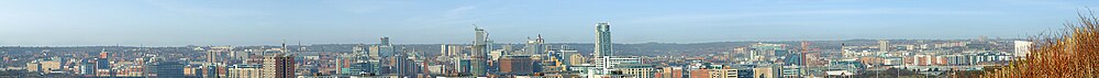 A panoramic view of a number of mainly modern high-rise buildings, some not yet complete, with, in the background, rising ground with suburban buildings and parks. The towers of some older buildings can be made out in the left foreground.