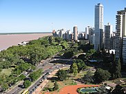 Photographie couleur. De hauts immeubles y surplombent une avenue et un square. L’avenue, empruntée par les automobiles, longe une étendue d’eau turbide. Ciel bleu dégagé.