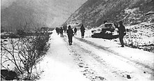 A line of soldiers walking past a destroyed tank