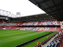 A stand from a football stadium with laid out turf and two adjacent goals in the foreground. On the claret stands are the words written in sky blue capitals "Sir Trevor Brooking stand" on top of the larger words "West Ham United". There are men in orange jackets scattered around the stadium.