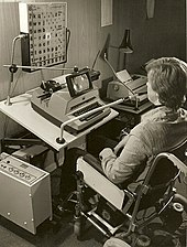 a black and white photograph showing a user sitting in front of a modified typewrite, there is grid of letters above the typewriter that appear to be lighting up in sequence, a tube goes from the users mouth to the machine