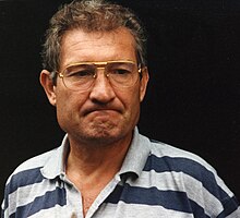 Photographie du buste d'un homme vêtu d'un polo rayé gris et bleu et portant des lunettes.