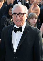 Headshot of a Caucasian male with thick rimmed glasses. He is wearing a black suit over a white shirt with a black bowtie.